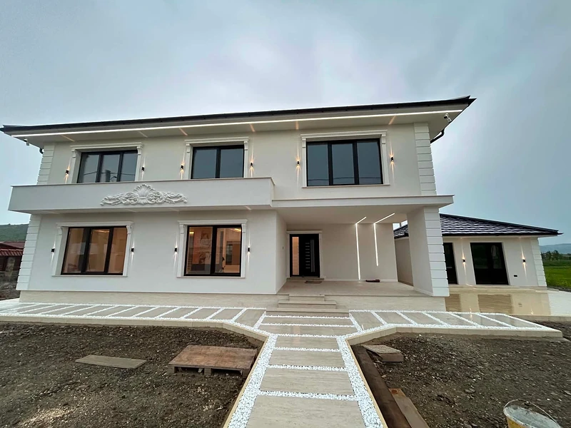 Casa construită, văzută frontal în lumina serii: fațadă albă cu tâmplărie neagră, aplice aprinse pe zid și pe conturul streșinii, iar la intrare aleea de dale cu pietriș alb.