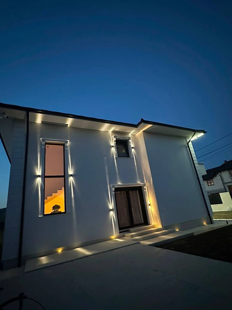 Casa seara, cu iluminatul exterior aprins pe zid și pe conturul streșinii — cadrul care se compară direct cu randarea de mai sus.